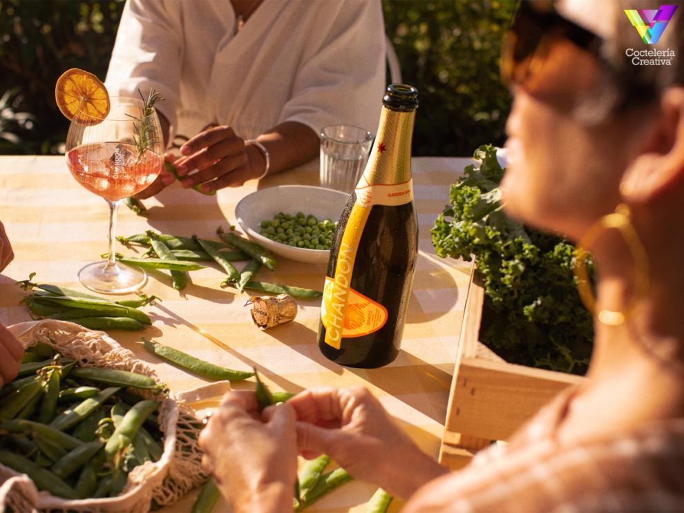 Mesa en el exterior con judías verdes y verduras, una copa de Chandon Garden Spritz con rodaja de naranja y una botella Mesa en el exterior con judías verdes y verduras, una copa de Chandon Garden Spritz con rodaja de naranja y una botella