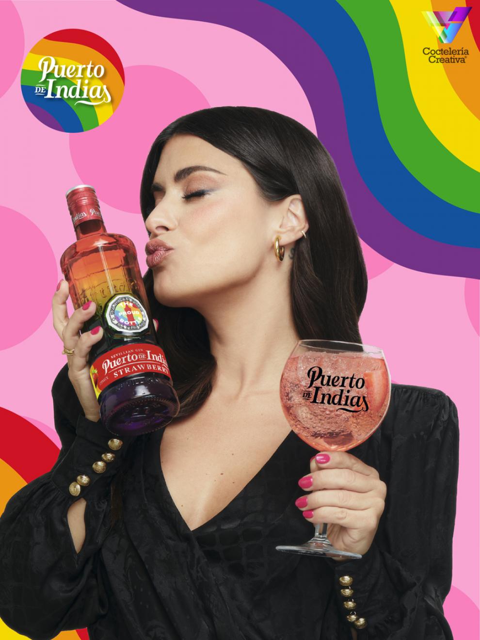 Mujer besando botella de Puerto de Indias con copa de Puerto de Indias en la mano y fondo con colores de la bandera LGBTIQ+